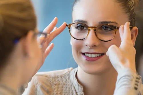 Junge Frau probiert eine Brille vor einem Spiegel an und lächelt dabei.