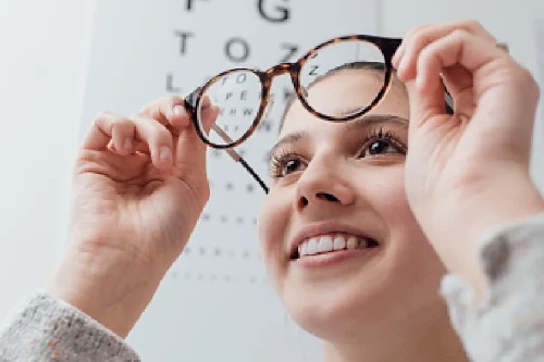Lächelnde junge Frau setzt ihre Brille auf; im Hintergrund ist eine Sehtafel zu erkennen.