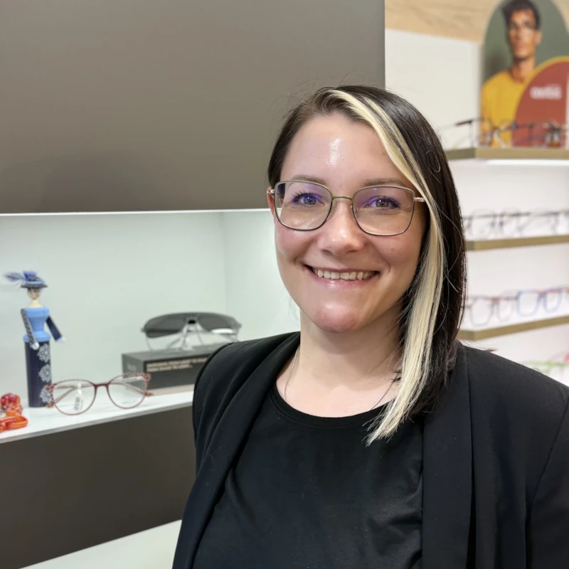 Lächelnde Optikerin mit Brille im Brillengeschäft vor Brillenregal